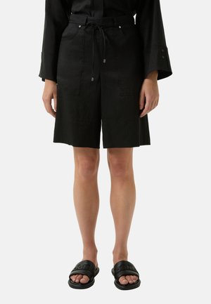 Schwarze Leinen-Shorts mit einem Kordelzugbund, zwei vorderen Taschen und lockerem Schnitt. Kombiniert mit flachen schwarzen Rutsch-Sandalen.