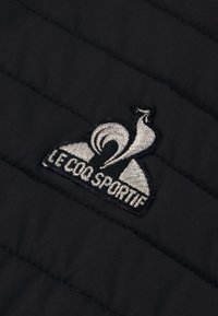 Tessuto di giacca nera trapuntata con un logo ricamato argentato che rappresenta un uccello e il testo "LE COQ SPORTIF" in basso.