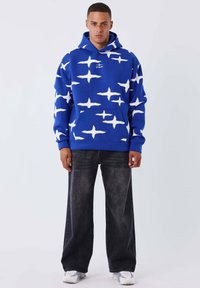 Sudadera azul con patrones abstractos blancos, bolsillo canguro frontal, capucha con cordón, combinada con jeans anchos oscuros y zapatillas blancas.