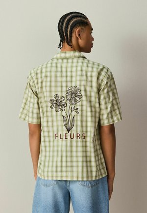 Personne aux cheveux tressés portant une chemise à manches courtes à carreaux verts avec une broderie florale noire et le texte "FLEURS" au dos.