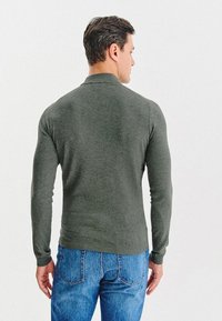 Zielony, dopasowany sweter wykonany z miękkiej dzianiny, z okrągłym dekoltem i ściągaczami, w zestawie z niebieskimi dżinsami.