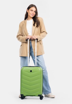 Valise rigide verte avec un design en chevron, poignées rétractables en argent et quatre roues noires. Fond blanc.