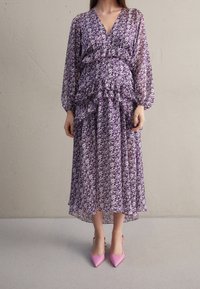 Robe à motifs floraux violets avec taille à volants et manches longues. Tissu transparent, longueur cheville avec une silhouette fluide, associée à des talons roses.