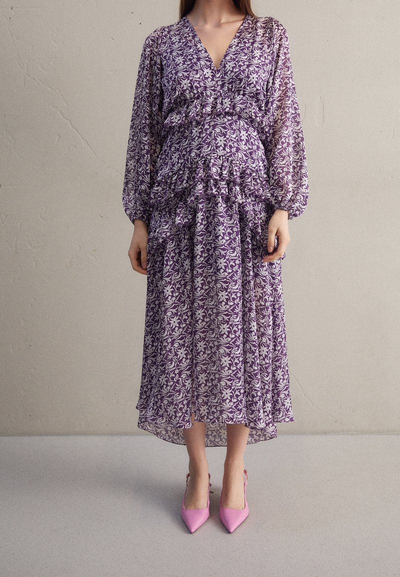 Robe à motifs floraux violets avec taille à volants et manches longues. Tissu transparent, longueur cheville avec une silhouette fluide, associée à des talons roses.