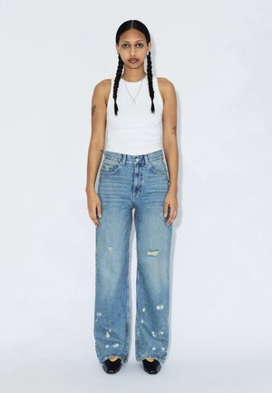 Jonge vrouw met twee lange vlechten, draagt een wit mouwloos topje, versleten wijde jeans en zwarte puntige schoenen, staand tegen een witte achtergrond.