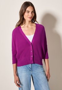 Paarse gebreide cardigan met een diepe V-hals, knopen aan de voorkant en driekwartmouwen, gecombineerd met lichtblauwe denim jeans.
