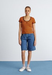 T-shirt à manches courtes rayé orange et marron associé à un short en denim bleu jusqu'aux genoux. Le modèle porte des ballerines blanches et des chaussettes gris clair.