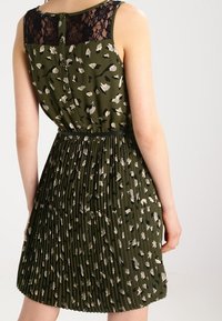 Vestido plisado verde con un respaldo de encaje negro y detalle de botones, con un patrón en beige y negro por todo el vestido.