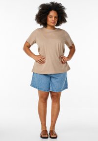 T-shirt beige à manches courtes avec un design texturé, associé à un short en denim bleu clair et des sandales marron. Modèle debout, les mains sur les hanches.