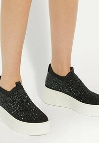 Des chaussures slip-on noires en tricot avec un dessus texturé ornés d accents scintillants, une semelle blanche épaisse et un col côtelé pour le soutien.