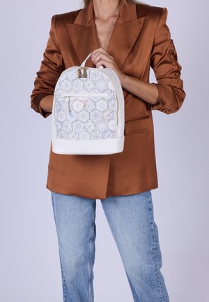 Persona che indossa un blazer marrone e jeans blu che tiene uno zaino bianco con motivo, tasca frontale con cerniera e dettagli dorati.