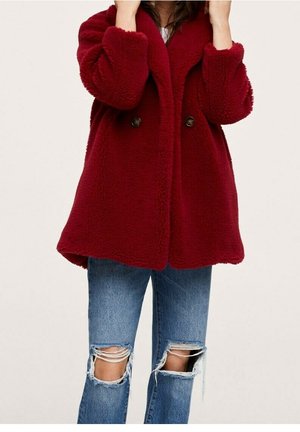 Czerwony płaszcz z polaru o dwurzędowym fasonie, z okrągłymi guzikami i luźnym krojem. Noszony na niebieskich, przetartych dżinsach z rozdarciami na kolanach.