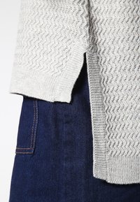 Ljust grå stickad tröja med ett zigzag-mönster, med en sidoklyfta vid fållen. Burit över mörk denim med synliga sömmar.