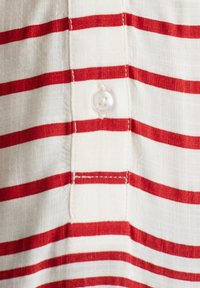 Tissu blanc à rayures horizontales rouges, avec une fermeture à boutons et une texture subtile. Détails de couture visibles le long des bords.