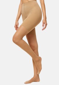 Frau in durchsichtigen, beigefarbenen, oberschenkellangen Formshorts mit passender transparenter Strumpfhose, die vor einem schlichten hellen Hintergrund steht.