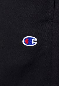 Petit logo brodé avec un "C" bleu et rouge sur un tissu noir texturé, probablement matière pour vêtements.