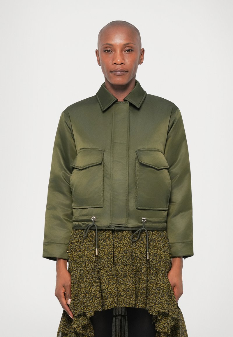 Haki zaļš īss bomber jakas satīna apdarē ar divām priekšējām kabatām ar atloku, regulējamu apakš malu un klasisku apkakli.