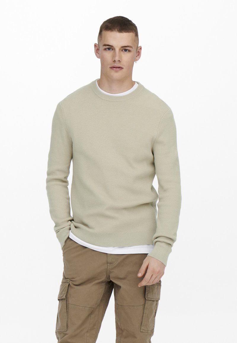 Suéter de canalé beige claro con mangas largas, escote redondo, usado sobre una camisa blanca; combinado con pantalones cargo color tostado que cuentan con bolsillos laterales.