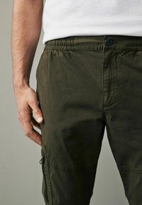 Olive žalios spalvos cargo kelnės su elastiniu juosmeniu, mygtukų užsegimu, šoninėmis kišenėmis ir užtrauktuko kišene ant šlaunies.