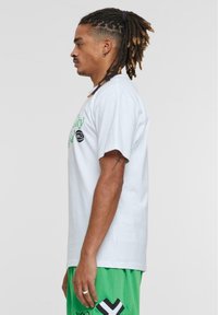 Hvid bomulds t-shirt med korte ærmer, med grøn tekst og et sort logo. Bæret med grønne mesh shorts, som viser en afslappet pasform.