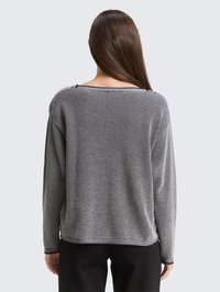 Donna con lunghi capelli castani che indossa un maglione grigio e nero con texture e pantaloni neri, vista da dietro su uno sfondo neutro.