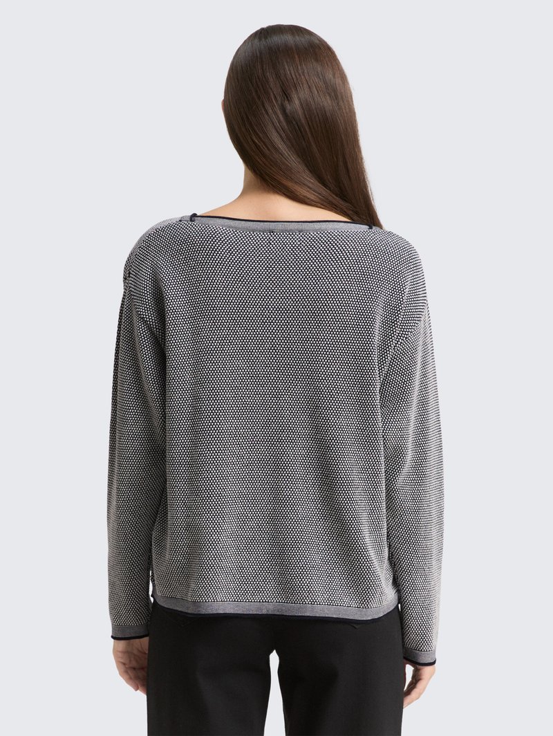 Kobieta z długimi brązowymi włosami, ubrana w szary i czarny sweter o fakturze oraz czarne spodnie, widoczna od tyłu na jednolitym tle.