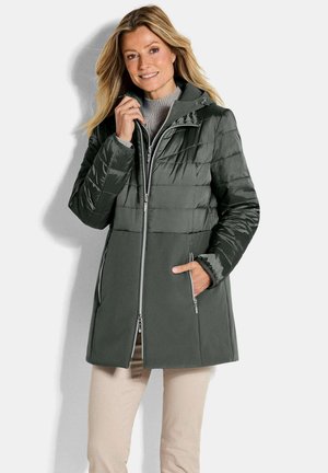 Femme portant un manteau d'hiver matelassé et en polaire vert foncé avec capuche, pantalon beige, souriante, debout sur fond blanc.