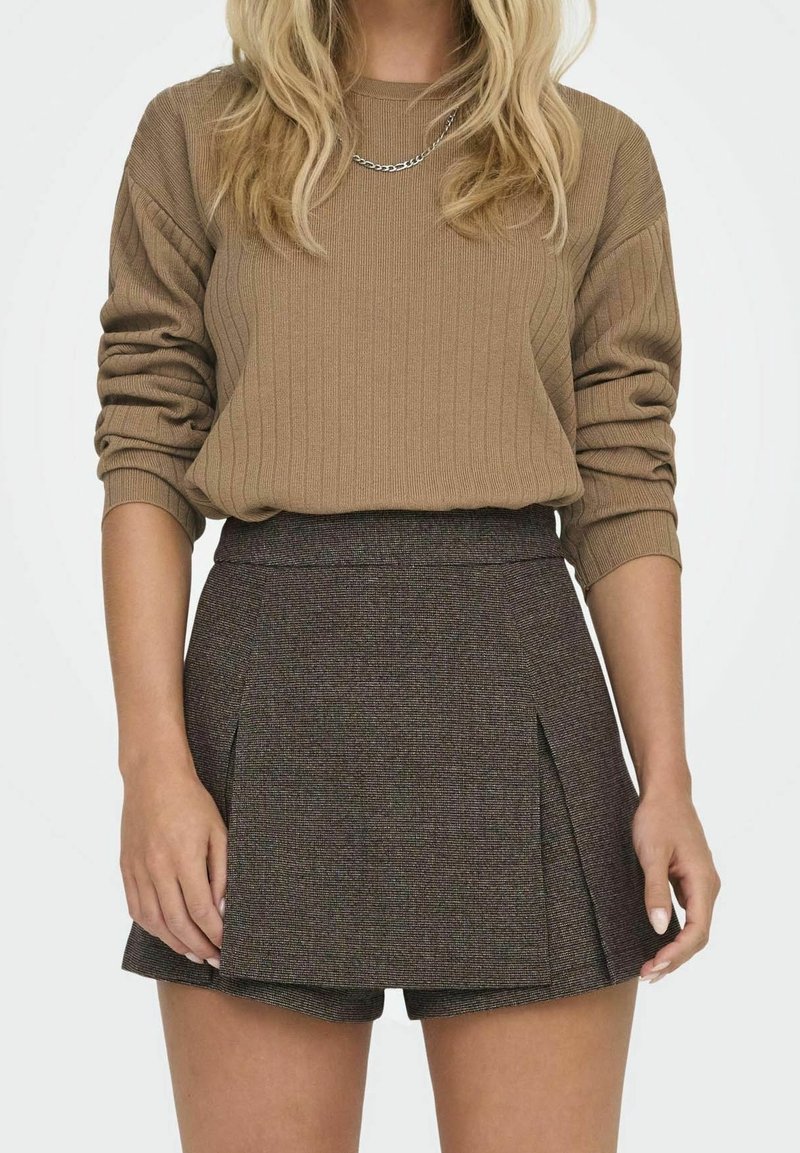 Femme portant un pull côtelé marron aux manches retroussées et une jupe plissée marron foncé avec un short intégré.