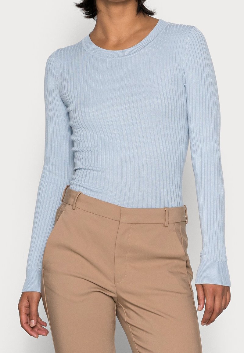 Femme portant un pull à manches longues côtelé bleu clair, rentré dans un pantalon beige taille haute avec passants de ceinture.