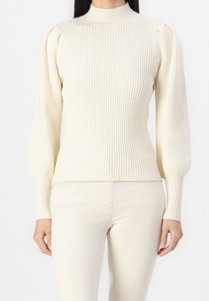 Femme portant un pull côtelé crème avec de longues manches bouffantes et un col montant, assorti d'un pantalon crème, debout devant un fond blanc.