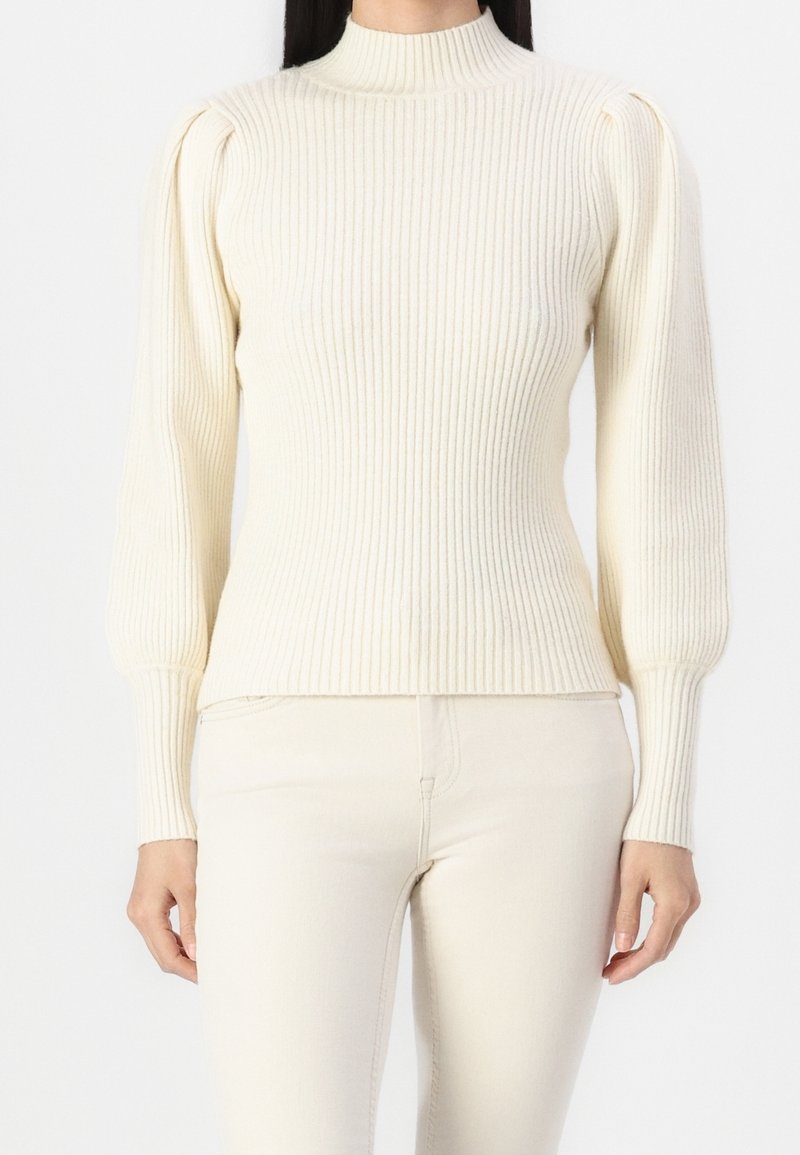 Femme portant un pull côtelé crème avec de longues manches bouffantes et un col montant, assorti d'un pantalon crème, debout devant un fond blanc.