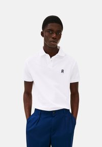 Polo shirt bianco a maniche corte con colletto, caratterizzato da un piccolo logo blu navy sul petto, abbinato a pantaloni su misura blu scuro.