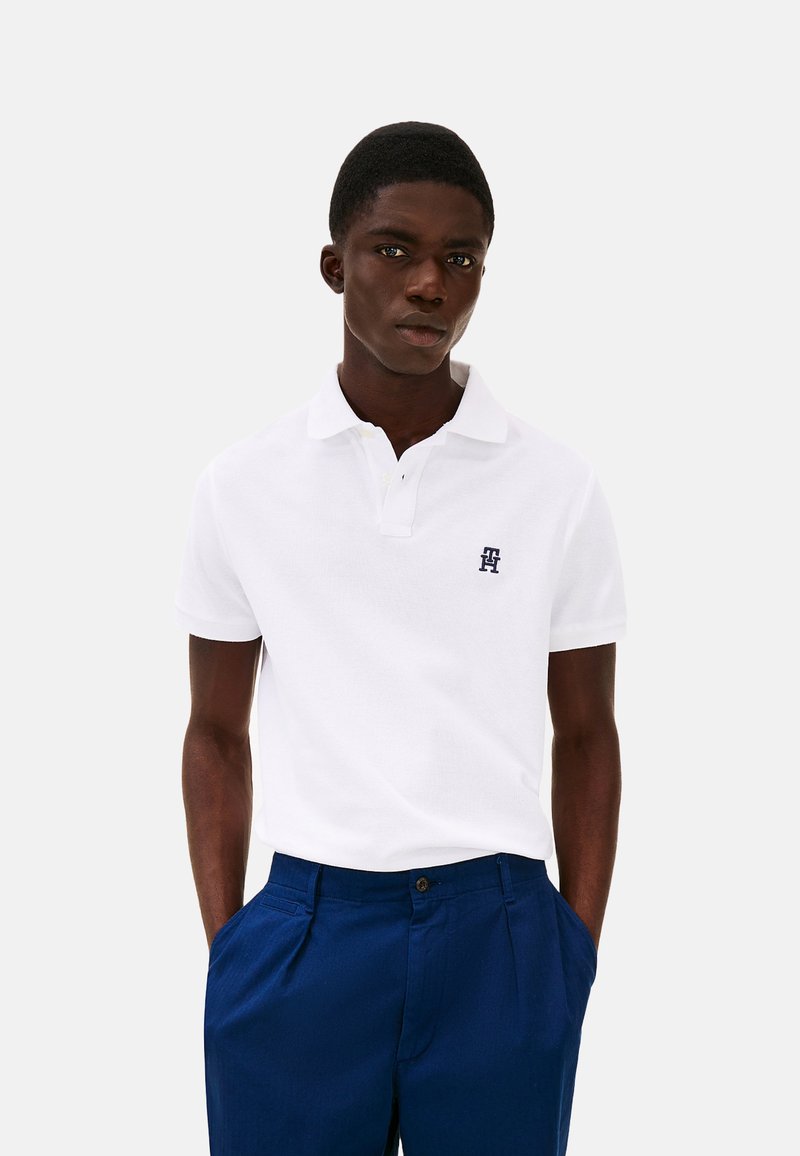 Polo shirt bianco a maniche corte con colletto, caratterizzato da un piccolo logo blu navy sul petto, abbinato a pantaloni su misura blu scuro.