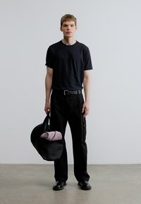 Fekete póló dombornyomott logóval, bő fekete nadrág, fekete cipő és egy nagy fekete táska, amelyben egy összehajtott lila tárgy található.