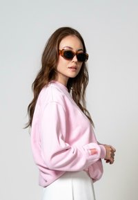 Rožinė megzta palaidinė su laisvu siluetu, įtempiamais rankogaliais ir siuvinėjimais. Modelis, vilkintis vėžlio kiauto akinius, stovi prieš šviesią foną.