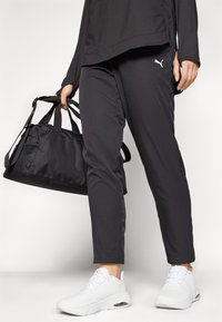 A person holds a black sports bag, wearing black pants and white athletic shoes. The outfit features a Puma logo.