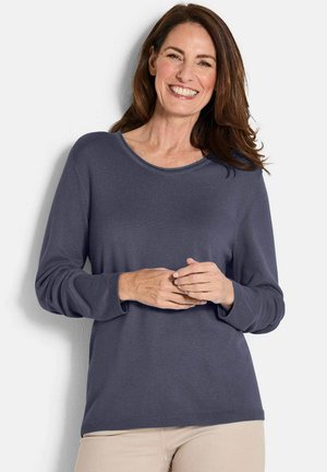 Glunderende vrouw met lang bruin haar, gekleed in een blauwe lange mouwen shirt en beige broek, staande tegen een witte achtergrond.