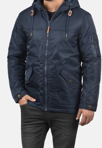 Uomo che indossa una giacca a cappuccio blu navy con zip frontale, tasche a patta e polsini regolabili, abbinata a jeans grigio scuro.
