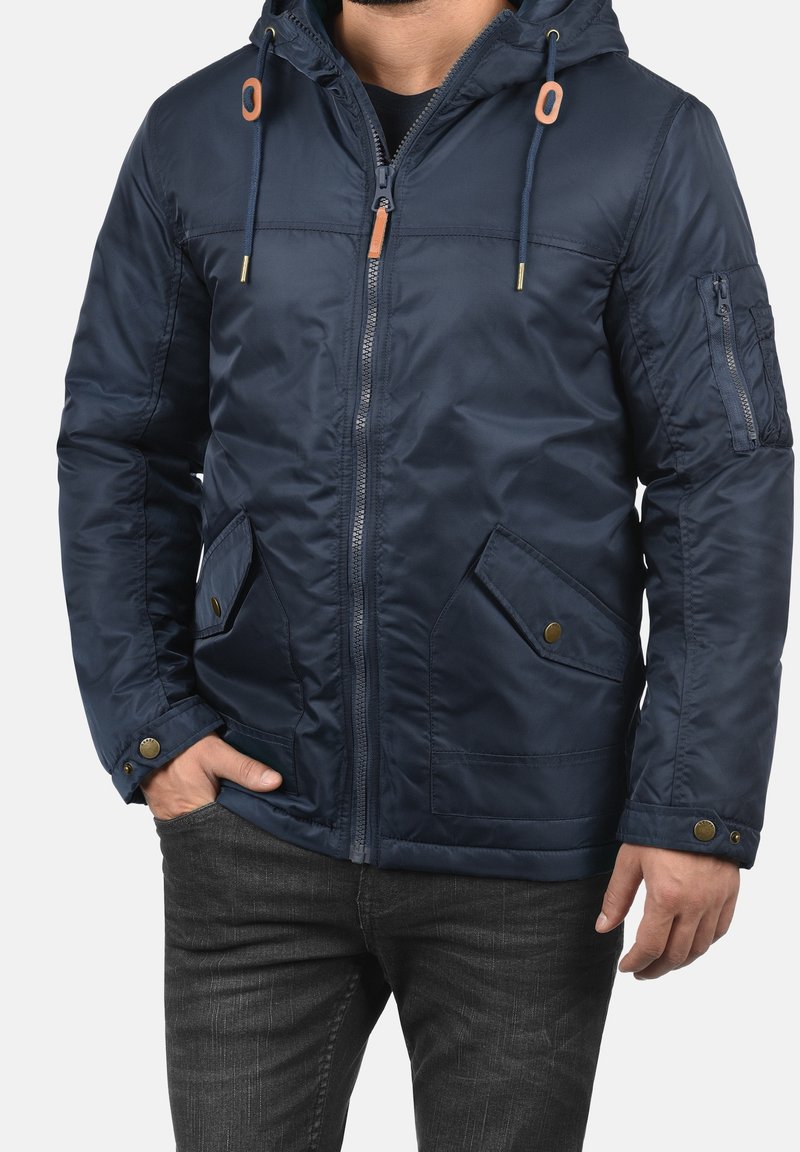Uomo che indossa una giacca a cappuccio blu navy con zip frontale, tasche a patta e polsini regolabili, abbinata a jeans grigio scuro.