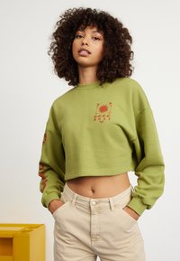Cropped green sweatshirt with red graphic design featuring sun and floral elements. Paired with light beige high-waisted pants.