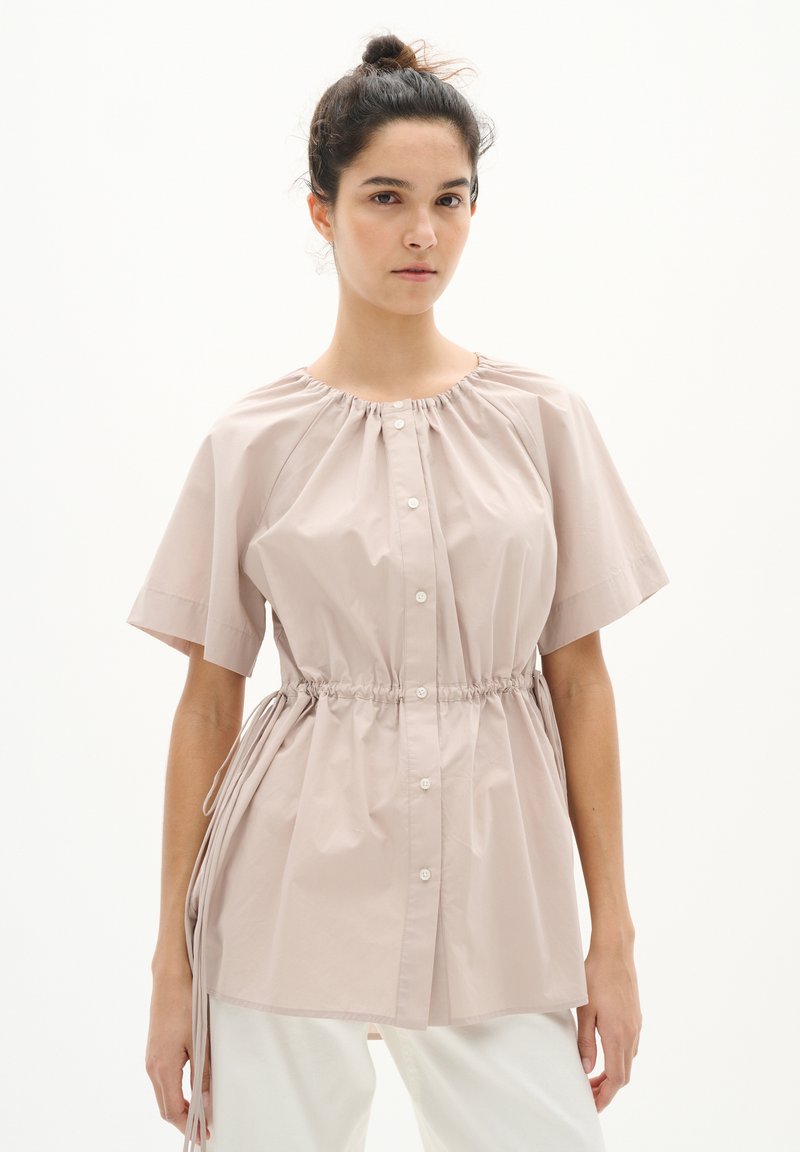 Blusa button-up rosa chiaro a maniche corte con scollatura arricciata, vita elastica e stringhe laterali. Realizzata in tessuto liscio.
