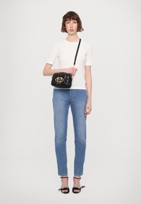 Borsa a tracolla in pelle nera trapuntata con logo dorato, indossata su una maglietta bianca semplice e jeans straight leg azzurri. Sandali neri con cinturini.