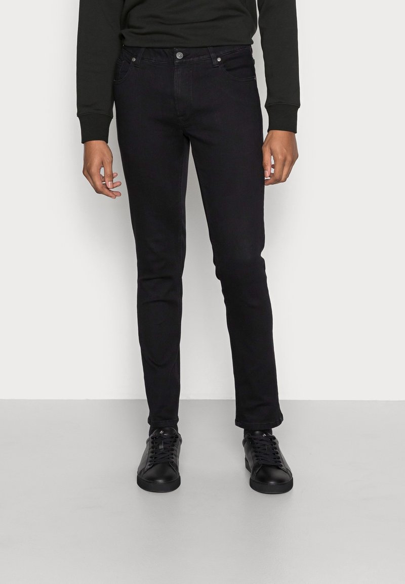 Homme portant un jean slim noir, un sweat-shirt noir et des baskets noires à lacets, debout sur un sol gris devant un mur blanc.