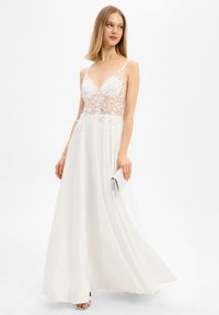 Vrouw die een witte avondjurk zonder mouwen draagt met kant en bloemmotieven op het lijfje, terwijl ze een zilveren clutch vast houdt.