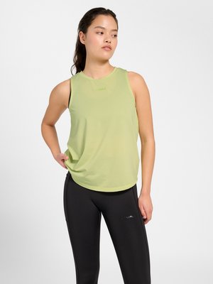 Kvinde med mørkt hår i hestehale iført ærmeløs lysegrøn top og sorte leggings, stående mod en hvid baggrund.