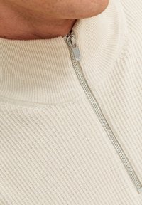 Krämfärgad stickad tröja med hög krage och silverfärgad dragkedja. Tyget har vertikala ribbor för textur och detalj.