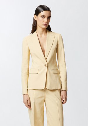 Blazer ajustado de color amarillo claro con un escote en V profundo, cierre de un solo botón, dos bolsillos frontales y mangas largas. Tejido suave y texturizado.