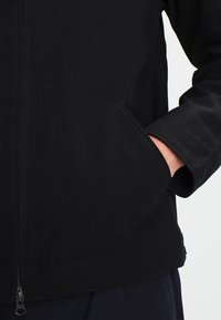 Černá plátěná bunda s kapsami na zip, bočními kapsami a pevným povrchem. Design zahrnuje rovný lem a dlouhé rukávy.