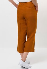 Pantalon court orange à texture lisse, avec une poche arrière et une coupe ajustée, associé à des baskets blanches.