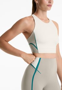 Kvinna som bär en beige och vit sportcrop-top och leggings med teal-accenter, drar i tyget vid midjan och visar klädernas elasticitet och passform.